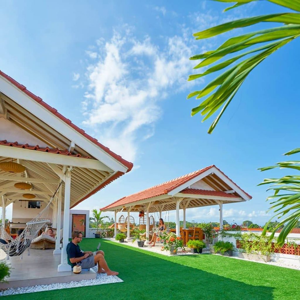 A rooftop coworking space with open buildings, plants, grass and people working from hammocks or swinging chairs.